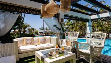 Zona junto a la piscina con mobiliario de mimbre, un sofá blanco y una mesa de vidrio con bebidas y un sombrero.