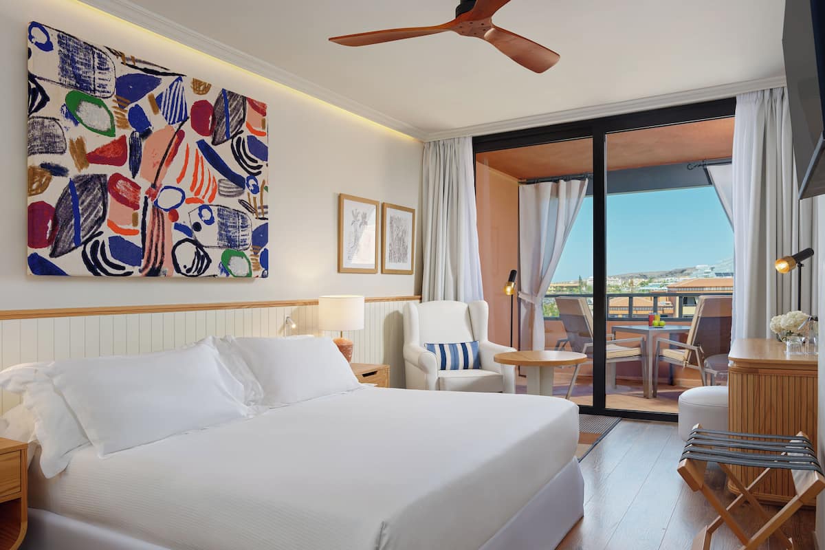 A hotel room with a large bed, a ceiling fan, a painting on the wall, a chair, a small table, and a view of the outside.