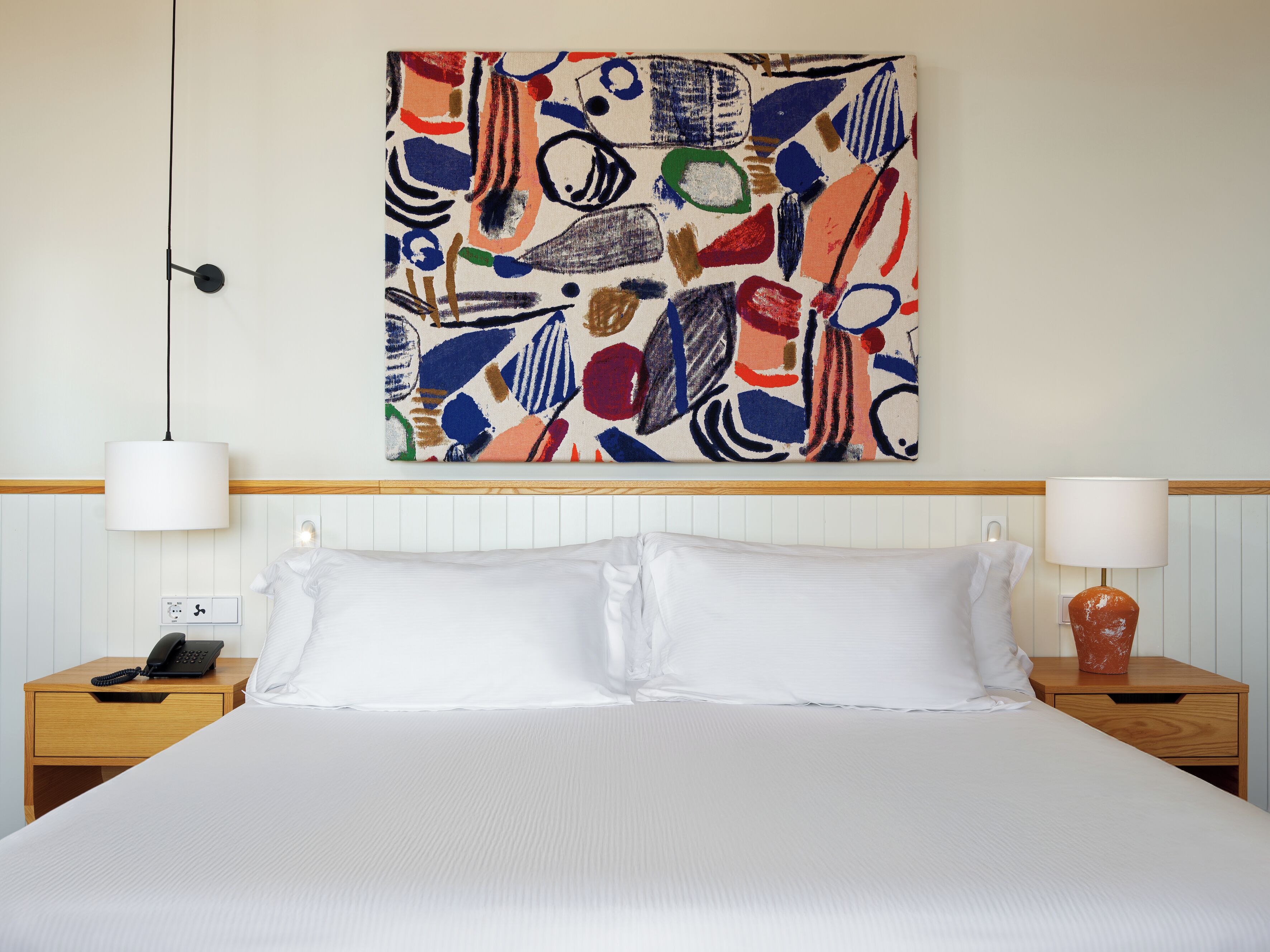 Une chambre d’hôtel avec un grand lit, du linge de lit blanc, une peinture abstraite aux couleurs vives accrochée au mur, une lampe de chevet et un téléphone sur la table de nuit.