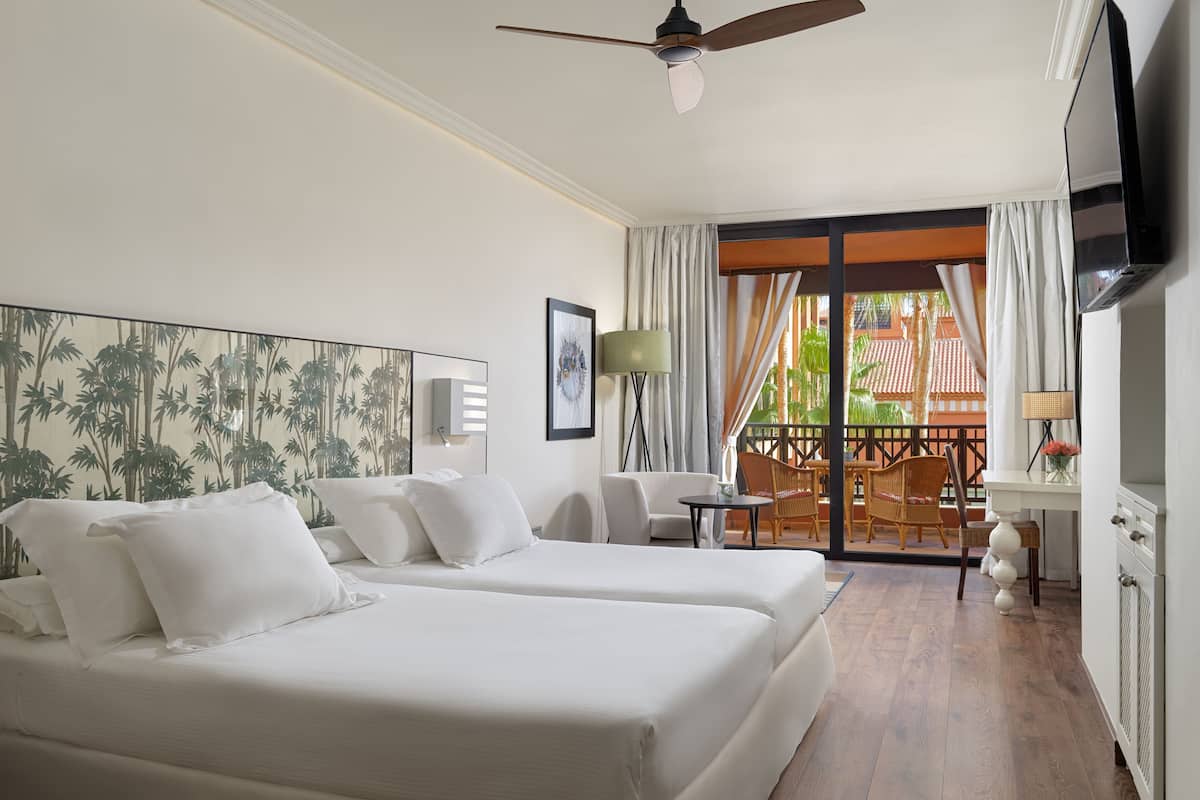A hotel room with two beds, a ceiling fan, a TV, a balcony with a view of palm trees, and a dining area with wicker furniture.