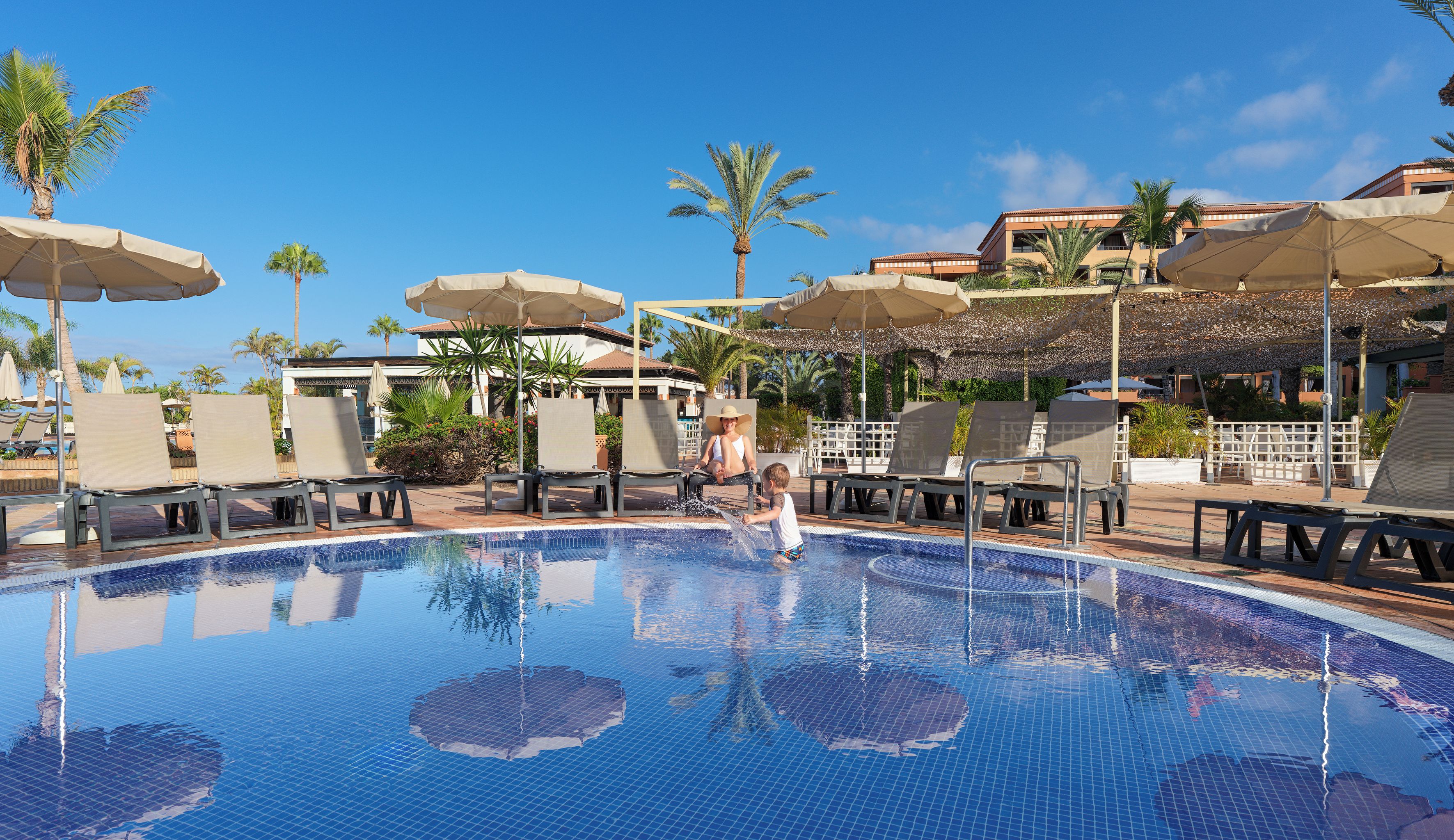 A swimming pool with lounge chairs and umbrellas, surrounded by palm trees and buildings.