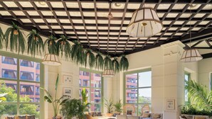 Amplia sala de estar con diversas opciones de asientos, incluyendo sofás y sillones, dispuestos alrededor de mesas bajas. El techo presenta un diseño de cuadrícula con plantas colgantes, y grandes ventanales ofrecen vistas de los edificios circundantes.
