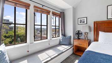 A bedroom with a large window, a wooden nightstand, a bed, and a view of the city.