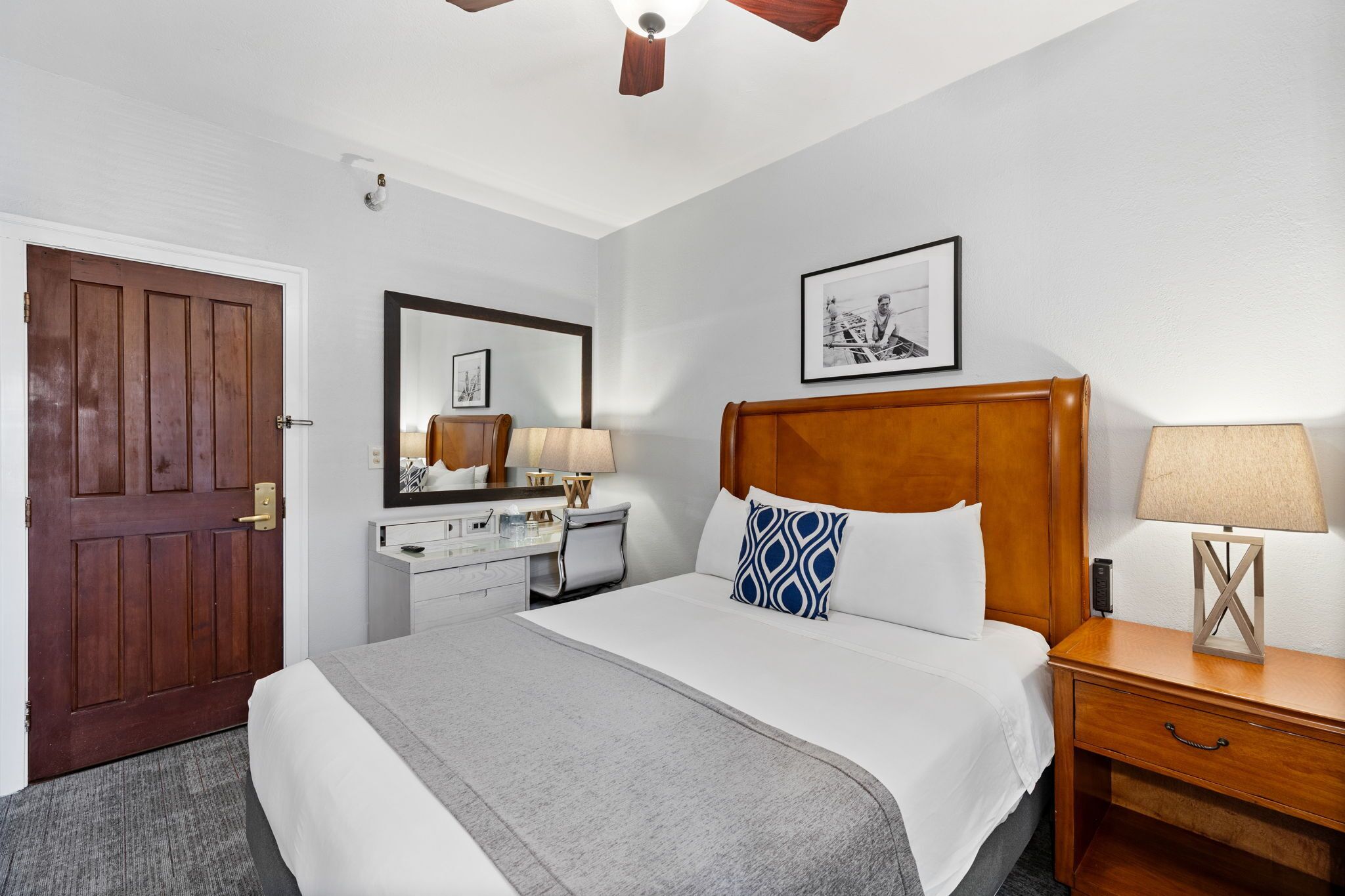 A hotel room with a large bed, a wooden headboard, a desk with a lamp, a dresser, and a door.