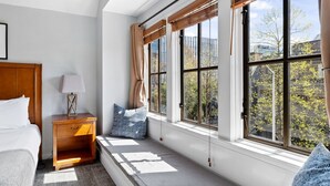 Un dormitorio con cama, mesita de noche, lámpara y vistas a árboles a través de amplias ventanas.