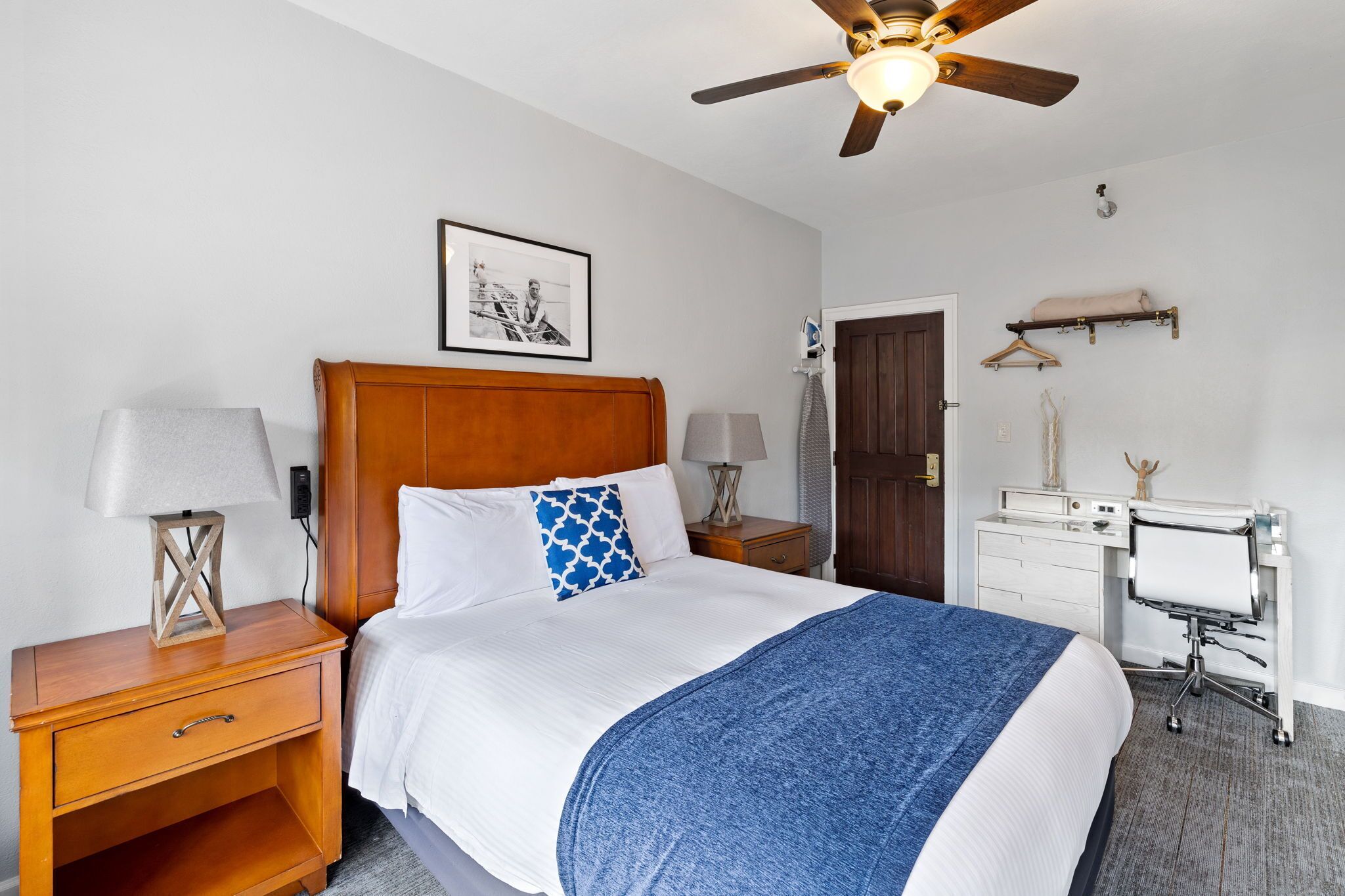 A neatly arranged hotel room with a wooden headboard bed, a bedside table with a lamp, a desk with a chair, and a ceiling fan.
