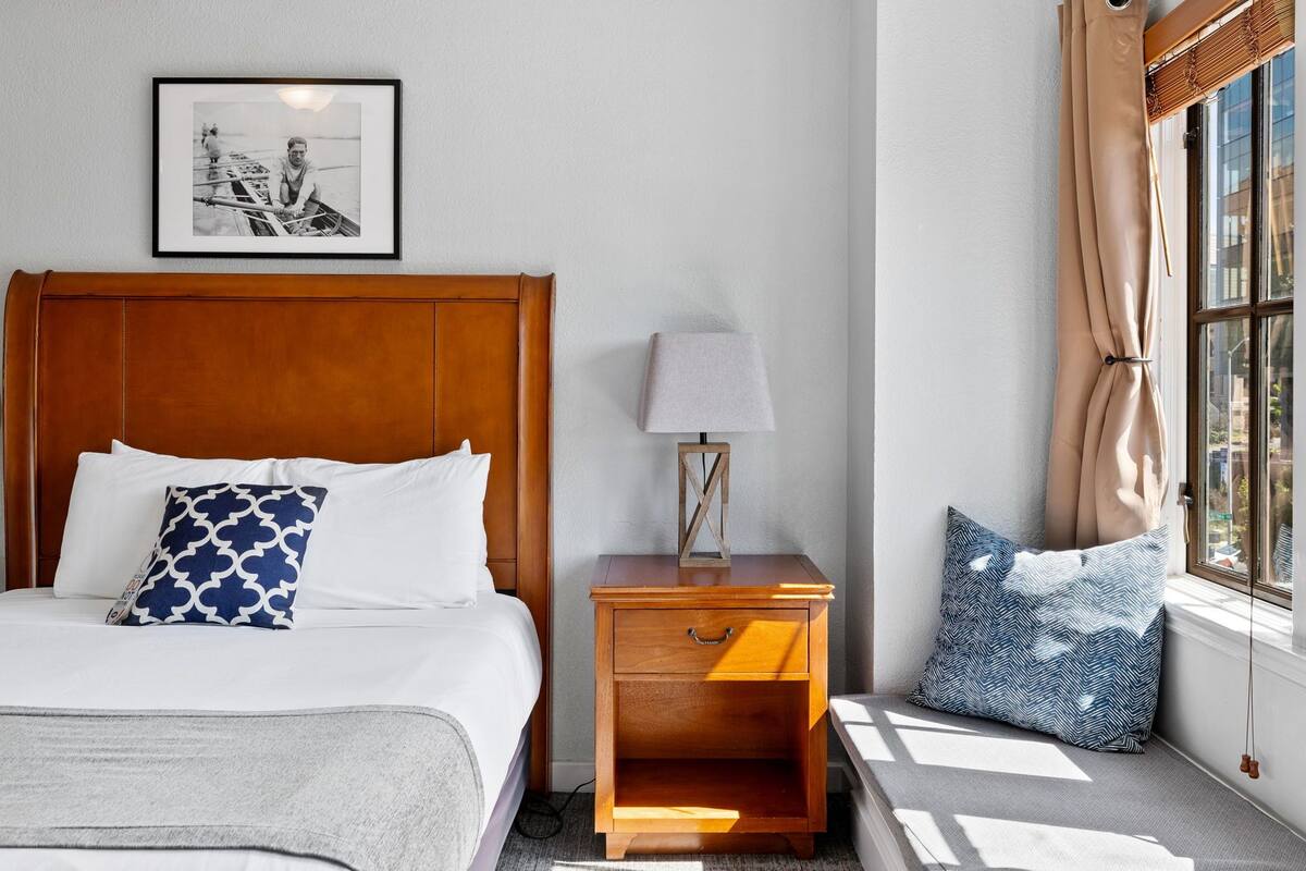 A neatly made bed with a patterned pillow, a bedside table with a lamp, and a window with curtains.