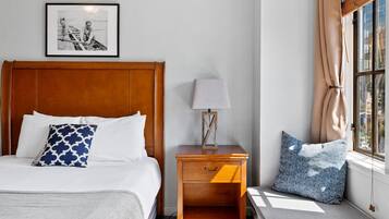 Una cama bien hecha con un cojín estampado, una mesita de noche con lámpara, y una ventana con cortinas.