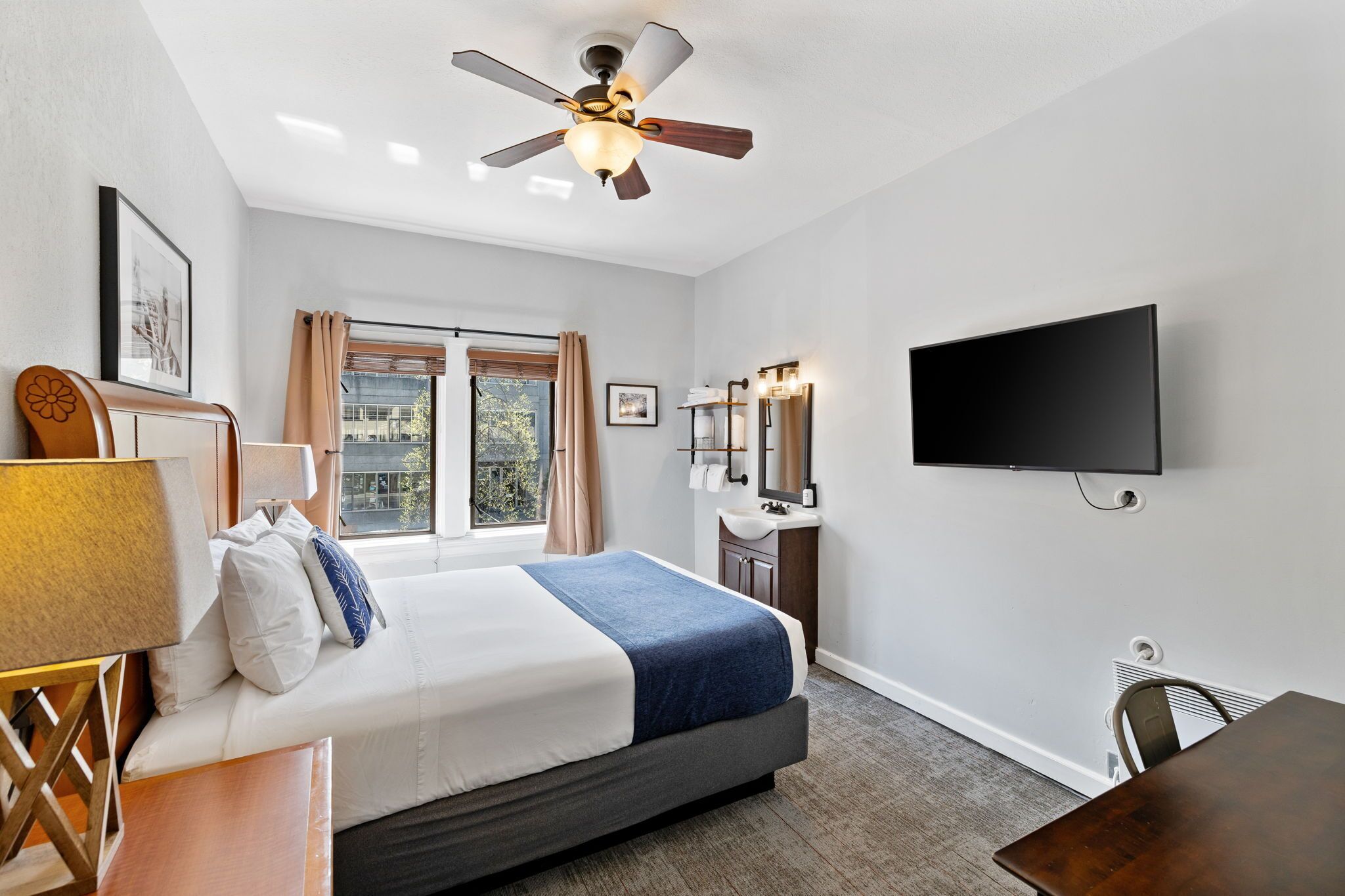 A hotel room with a bed, a desk, a chair, a television, a ceiling fan, a window with curtains, and a bathroom with a sink and mirror.
