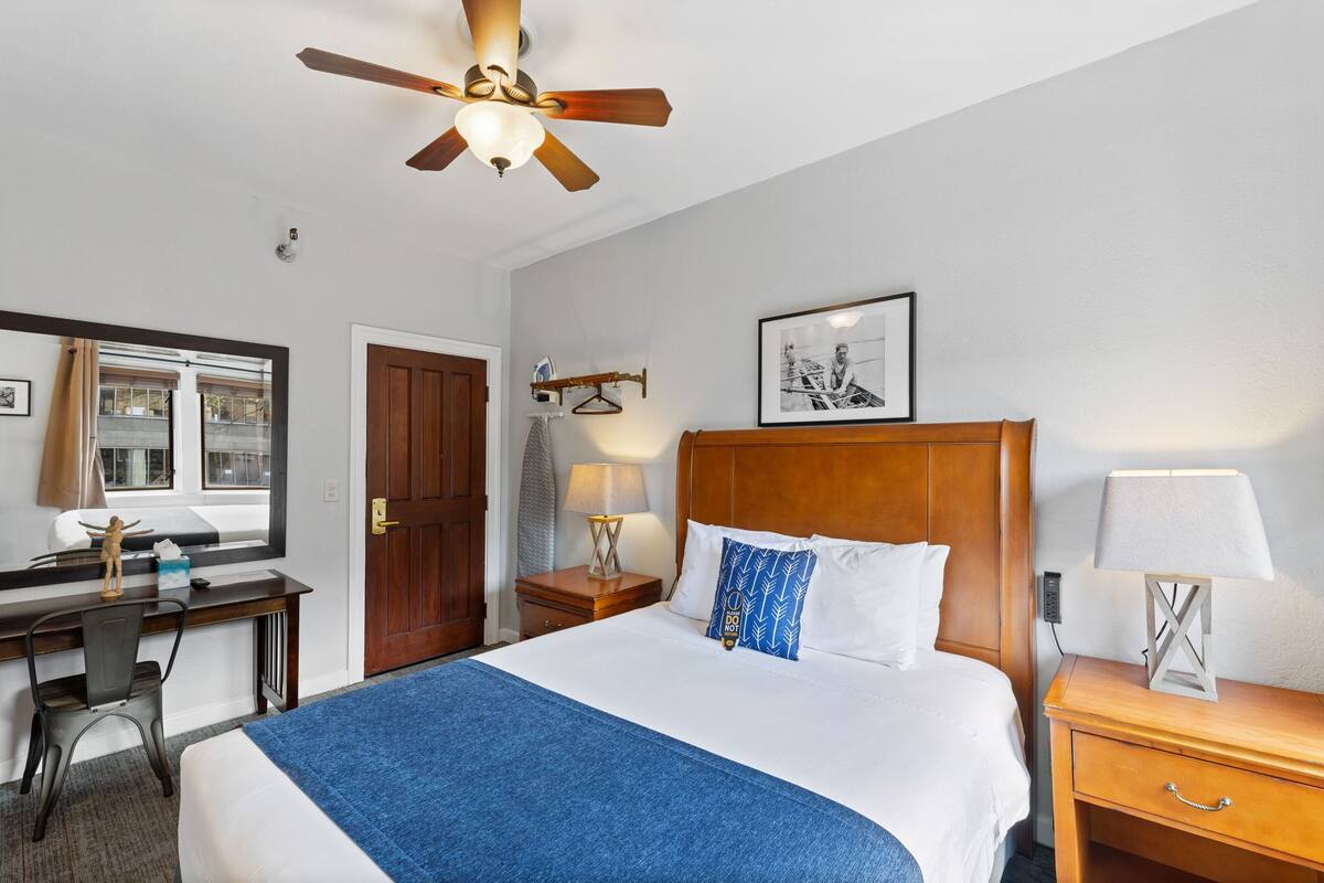 A hotel room with a bed, a desk, a chair, a bedside table, a lamp, a ceiling fan, a window, and a door.