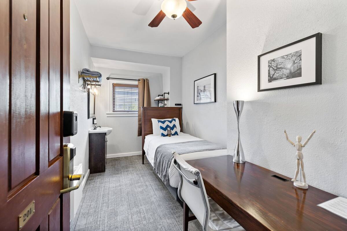 A hotel room with a bed, bedside table, desk, and a window with curtains.