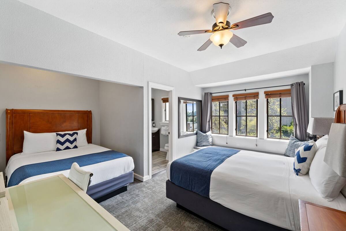 A hotel room with two beds, a ceiling fan, and a view of the outdoors through large windows.