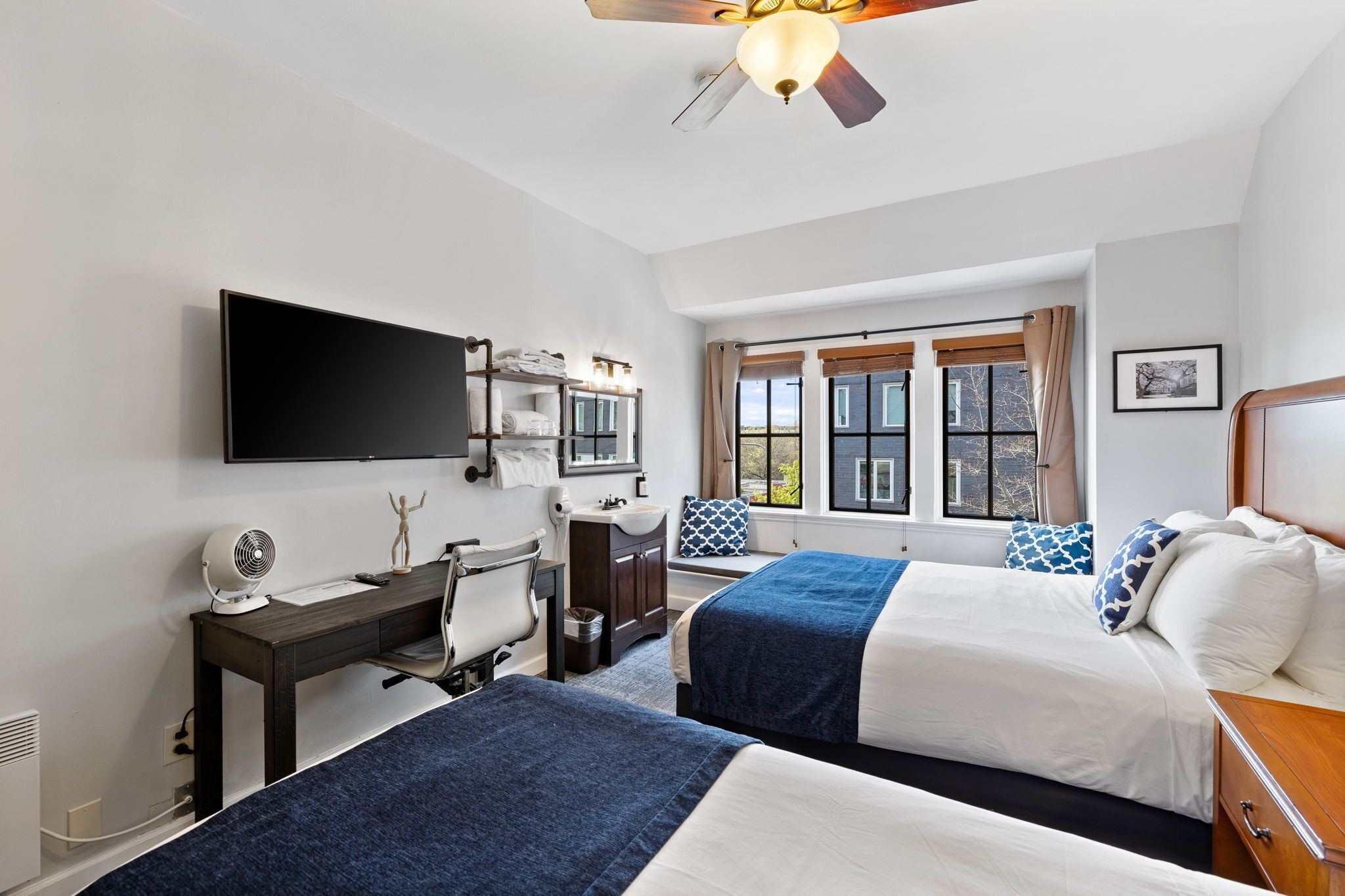 Ein Hotelzimmer mit zwei Betten, einem Schreibtisch mit Stuhl, einem Fernseher, einem Deckenventilator und einem Fenster mit Vorhängen.