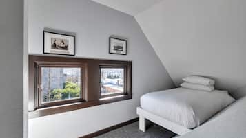 A bedroom with a sloped ceiling, a bed, a window with a view of buildings, and two framed pictures on the wall.
