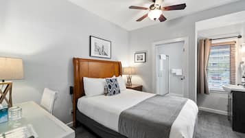 Habitación de hotel con cama, mesitas de noche, escritorio, ventilador de techo y ventana con persianas.