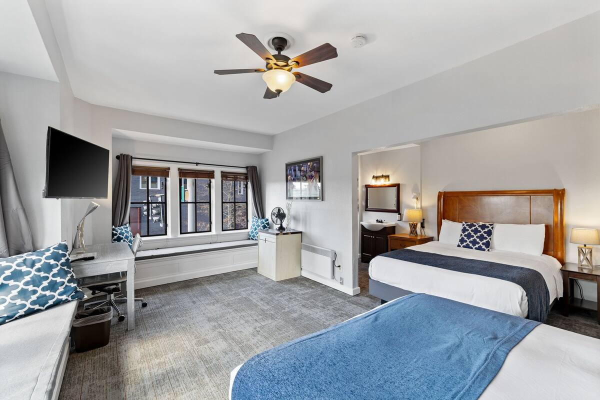 A hotel room with two beds, a desk, a TV, a ceiling fan, a window with curtains, a bathroom with a sink and mirror, and a radiator.