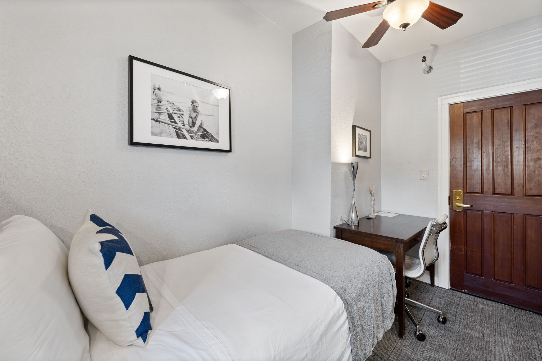 A bedroom with a bed, a desk, and a chair. There is a door and a ceiling fan.
