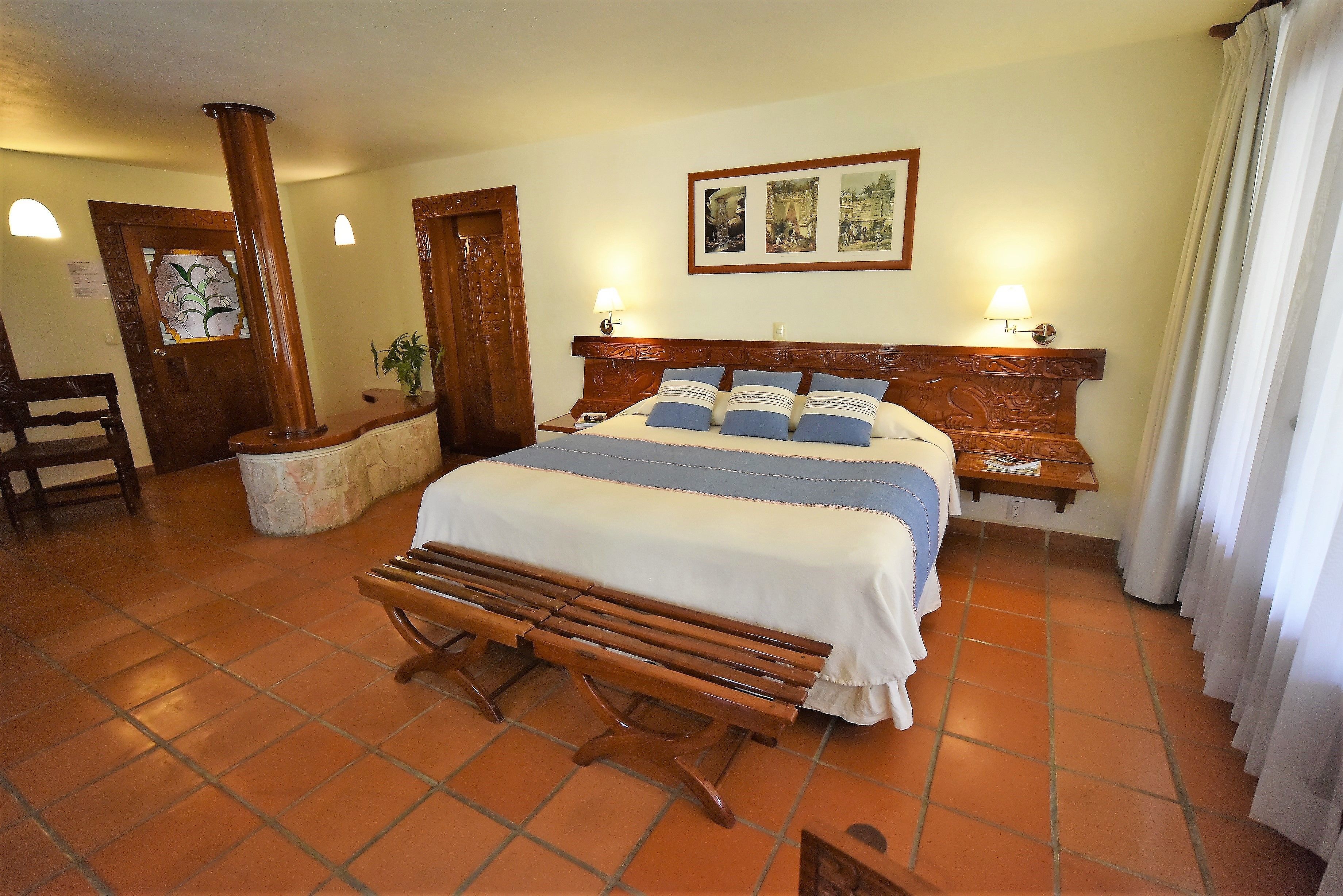 A hotel room with a large bed, wooden headboard, two bedside lamps, a bench, and a tiled floor.