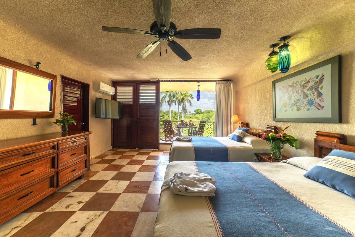 Una camera d'albergo con pavimento a scacchi, una ventola a soffitto, un letto con copriletto bianco e blu, un comò in legno, una TV e un balcone con vista su palme.