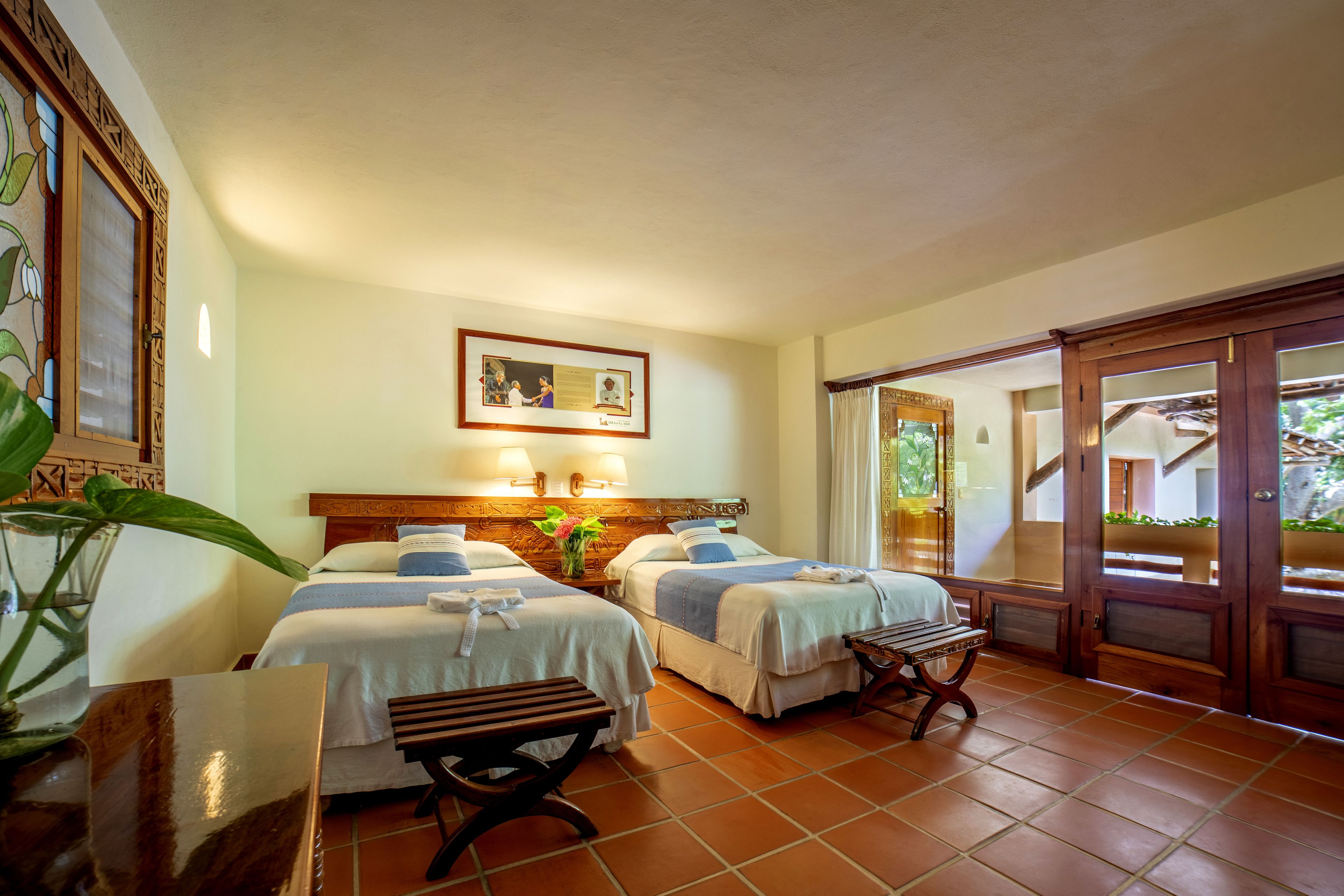 A hotel room with two beds, a wooden bench, a table, and a large window.