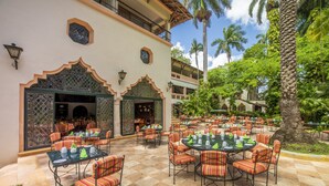 An outdoor dining area with round tables and striped chairs, surrounded by palm trees and a building with ornate arches. - Mayaland Hotel & Bungalows (Chichen Itza)