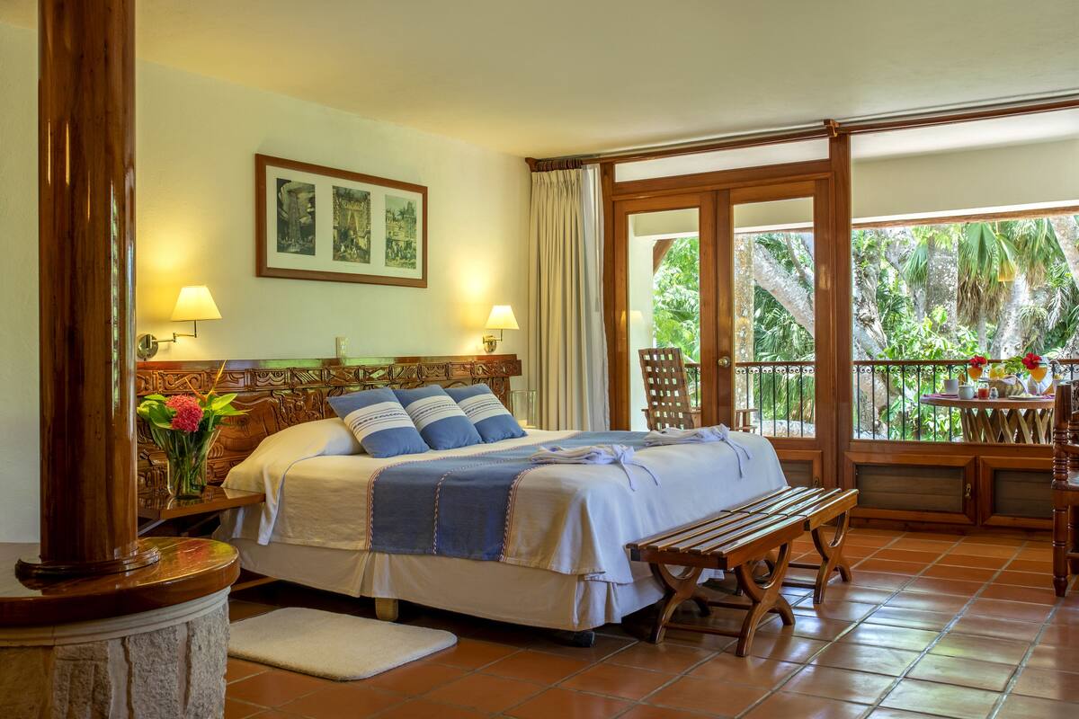 Camera da letto con un grande letto, testiera in legno, due lampade ai comodini, una panca e vista sul verde attraverso le porte del balcone.