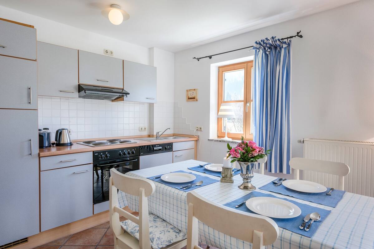 Une cuisine de taille modeste, avec des meubles blancs, une cuisinière et un évier. On y trouve une table à manger mise avec des assiettes, des couverts et un vase rempli de fleurs.