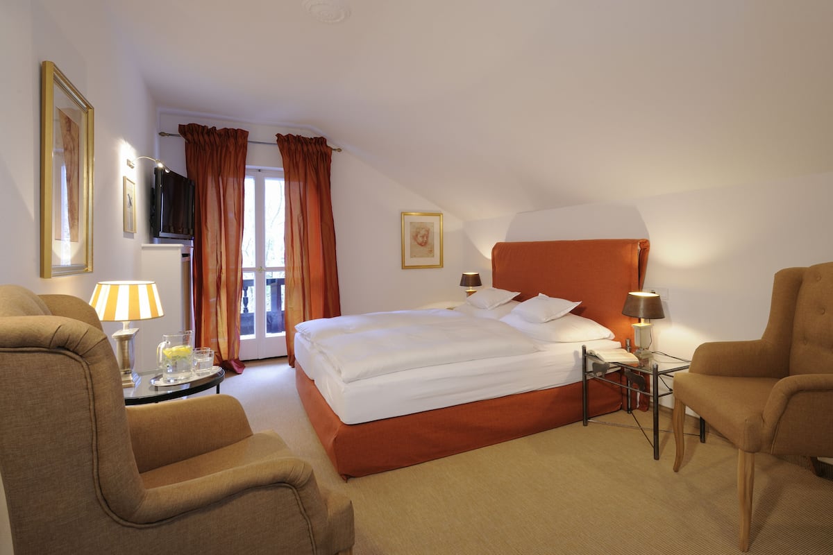 Une chambre d’hôtel comprenant un lit, deux fauteuils, une table de chevet avec une lampe, un téléviseur et une fenêtre avec des rideaux.