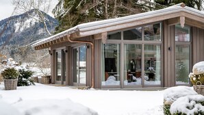 Ein modernes Chalet mit großen Glasfenstern, einer Holzfassade und Blick auf schneebedeckte Berge.