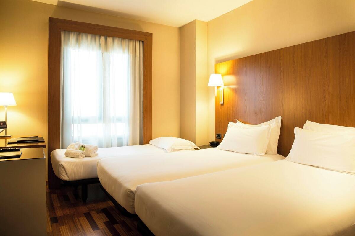 A hotel room with two beds, a desk, a chair, a lamp, and a window with curtains.