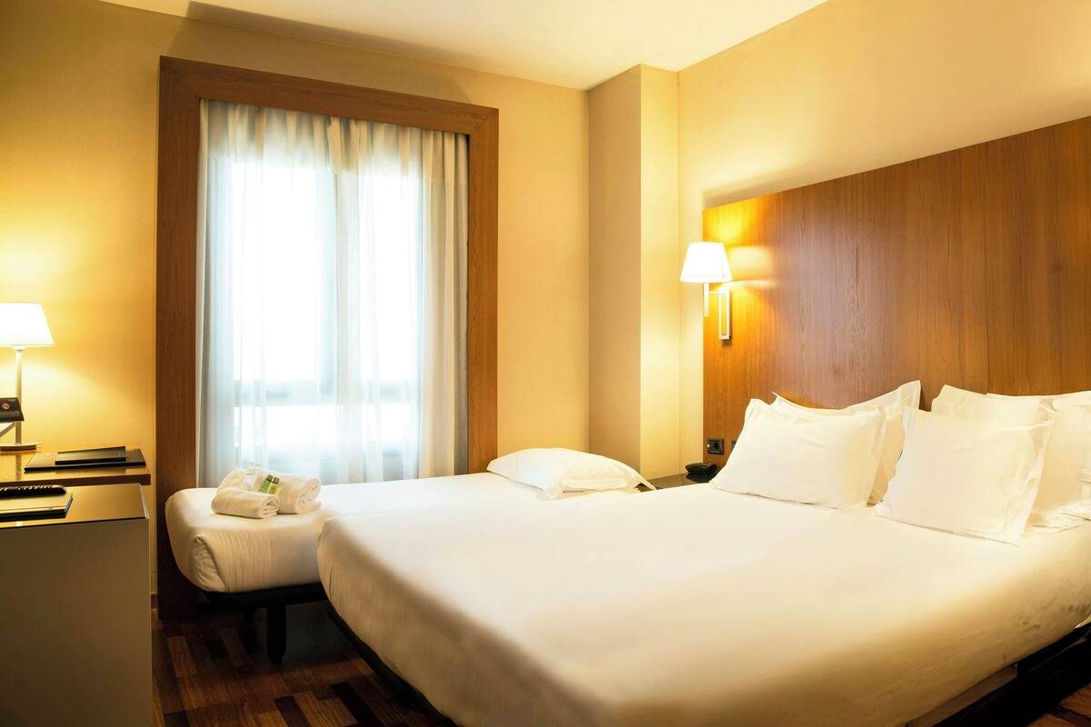 A hotel room with a large bed, a desk, a chair, a lamp, and a window with curtains.