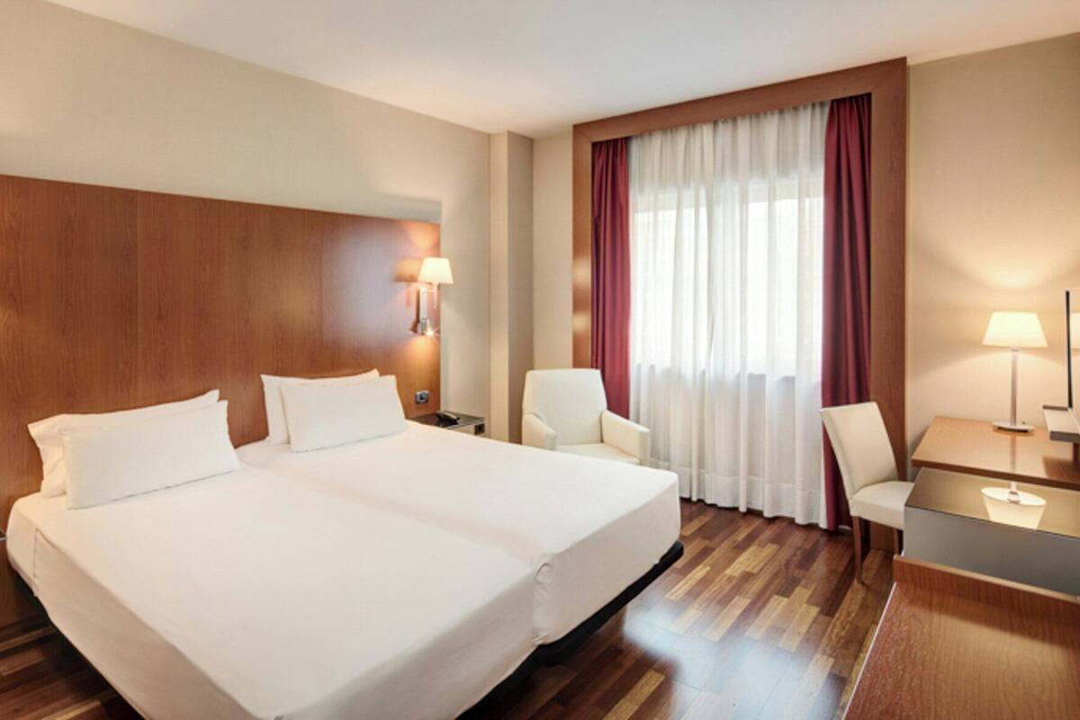 A hotel room with a large bed, two bedside tables, a desk with a chair, a TV, and a window with curtains.
