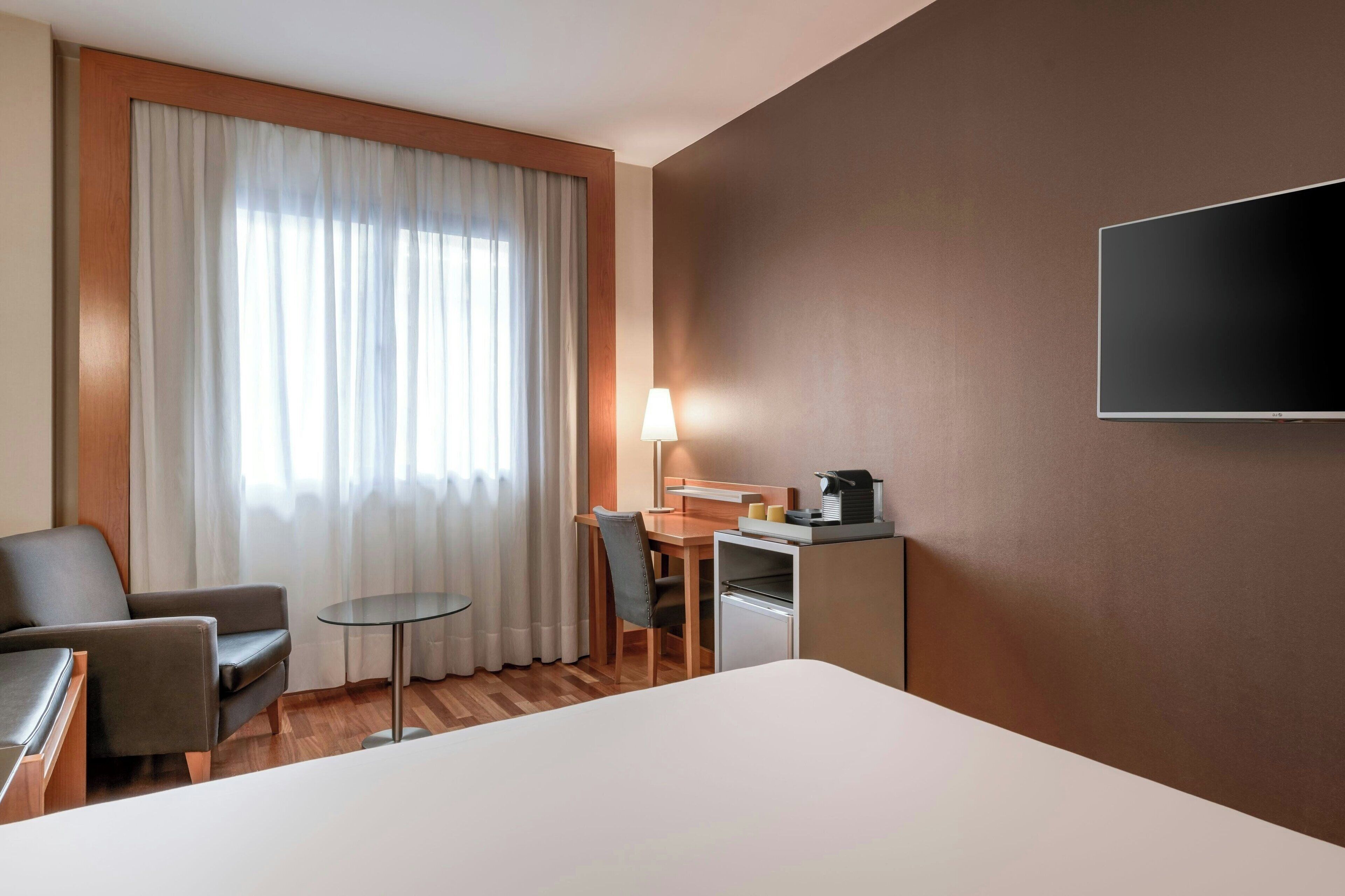 A hotel room with a bed, a desk, a chair, a lamp, and a TV.