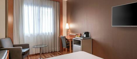 A hotel room with a bed, a desk, a chair, a lamp, and a TV.