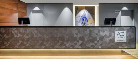 A hotel reception desk with a modern design, featuring a geometric patterned wall, a wooden accent wall, and a sign indicating the hotel name.