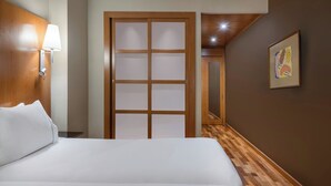 A hotel room with a large bed, a wooden door with frosted glass panels, a wall-mounted lamp, and a framed abstract painting.