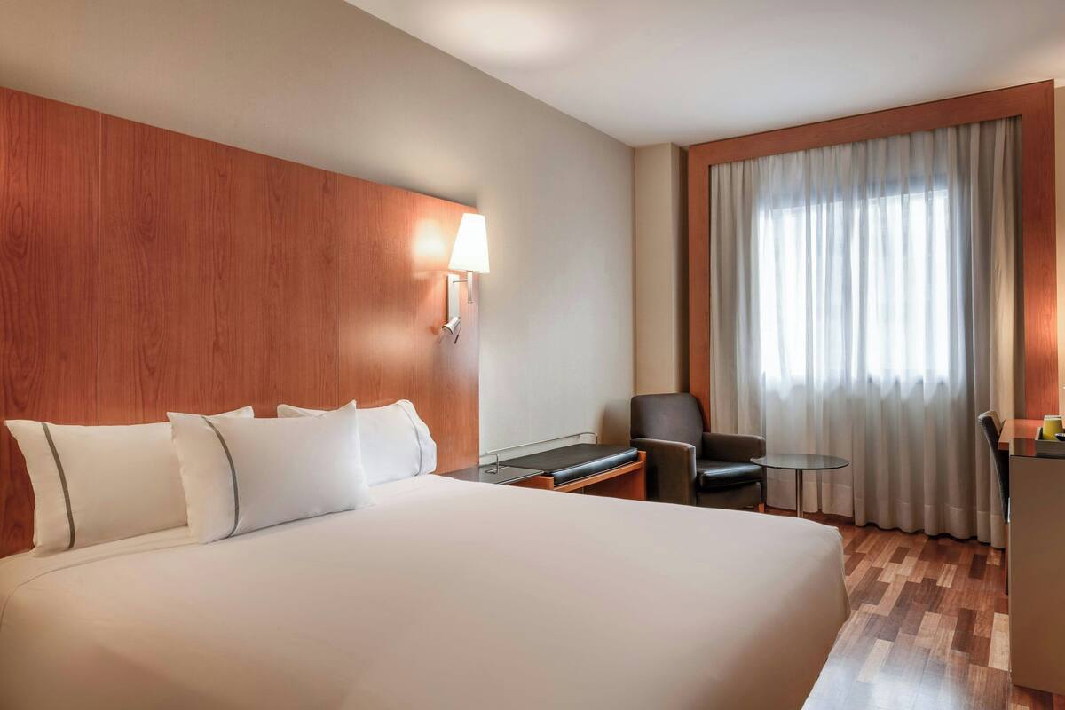 A hotel room with a large bed, a desk, a chair, a lamp, and a view of the outdoors through a window with curtains.