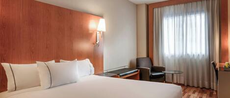 Una habitación de hotel con una cama grande, un escritorio, una silla, una lámpara y vistas al exterior a través de una ventana con cortinas.