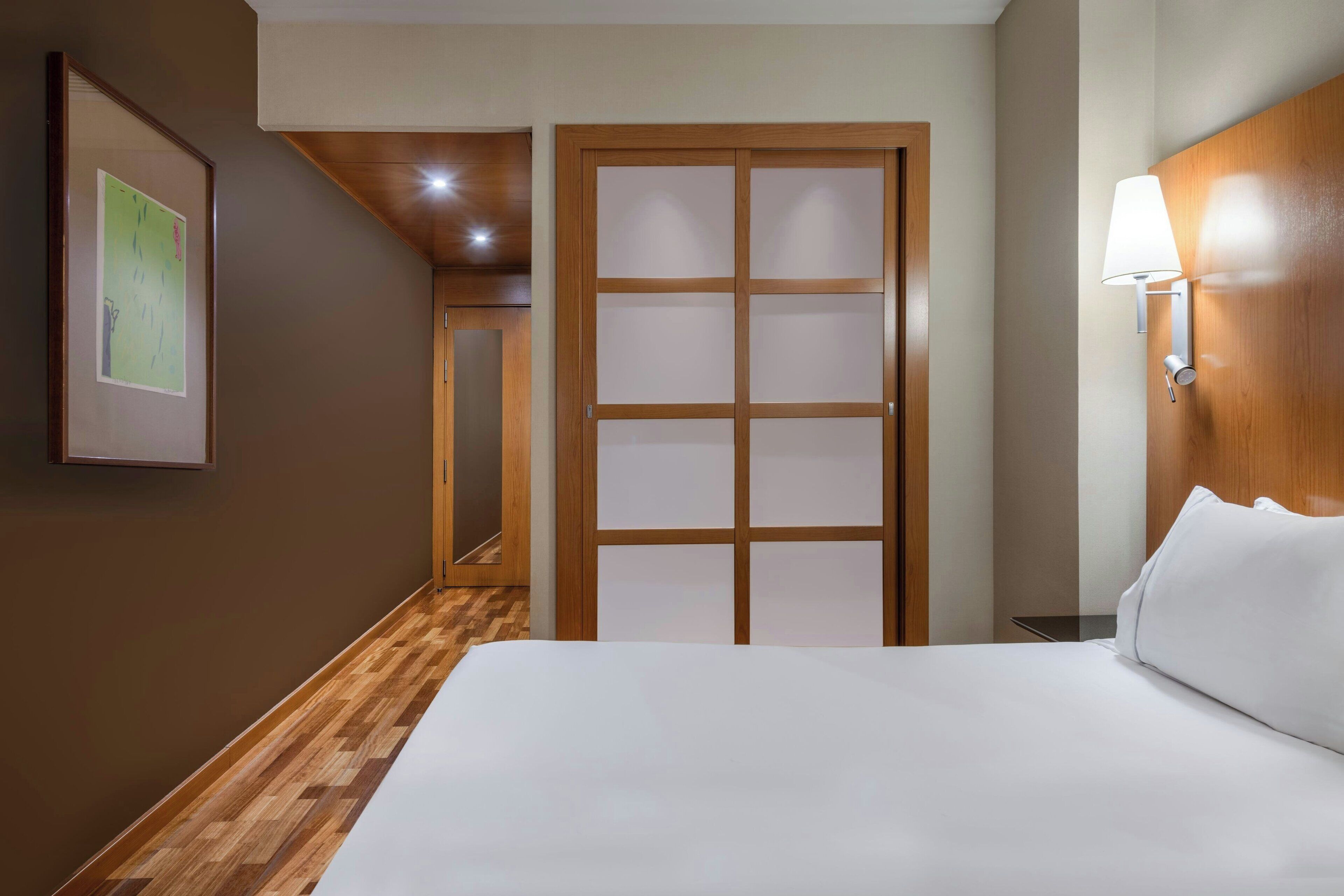 A hotel room with a wooden floor, a bed with white bedding, a wooden door with frosted glass panels, and a wall-mounted lamp.