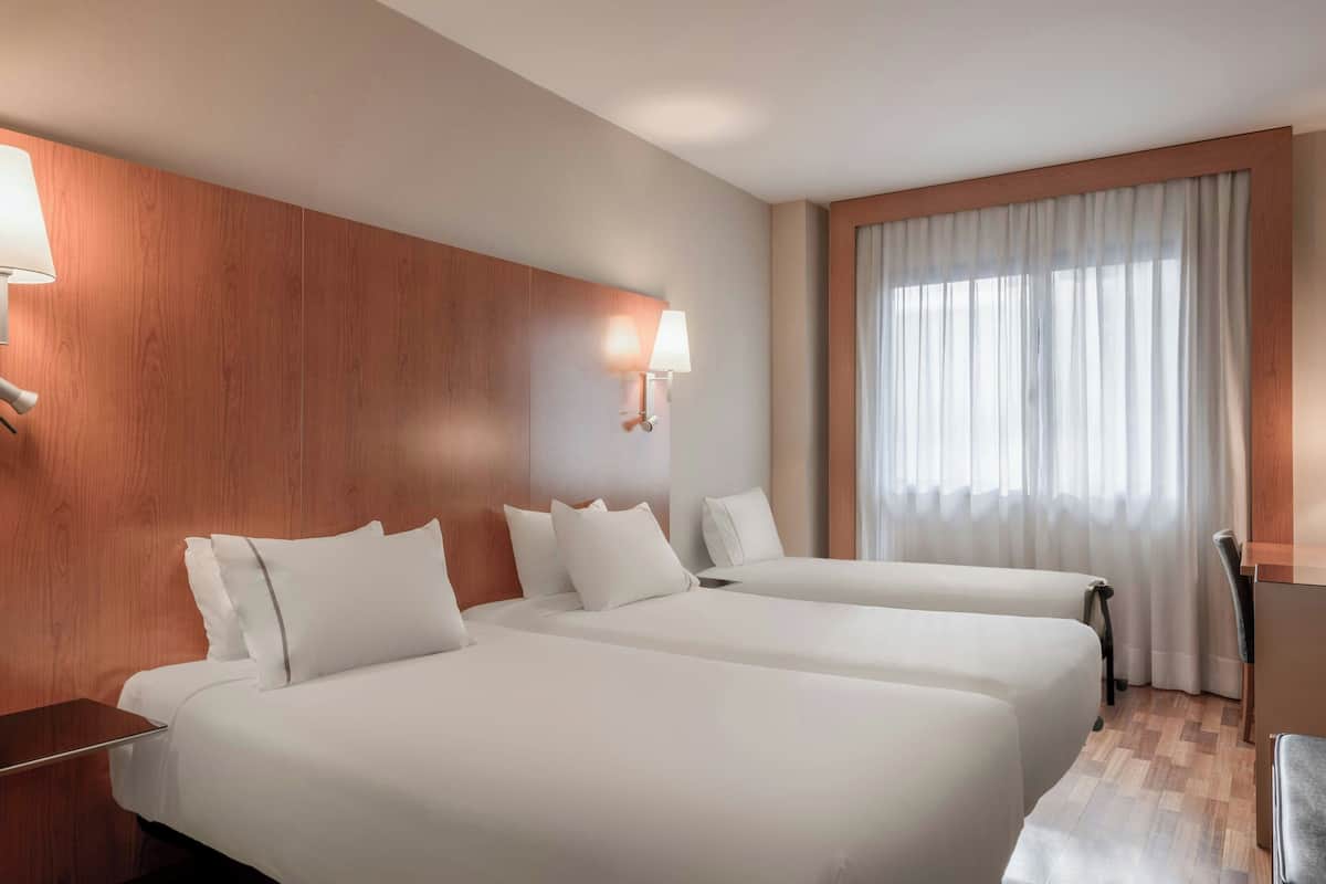 A hotel room with two beds, wooden paneling on the walls, a desk, and a window with curtains.