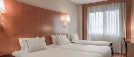A hotel room with two beds, wooden paneling on the walls, a desk, and a window with curtains.
