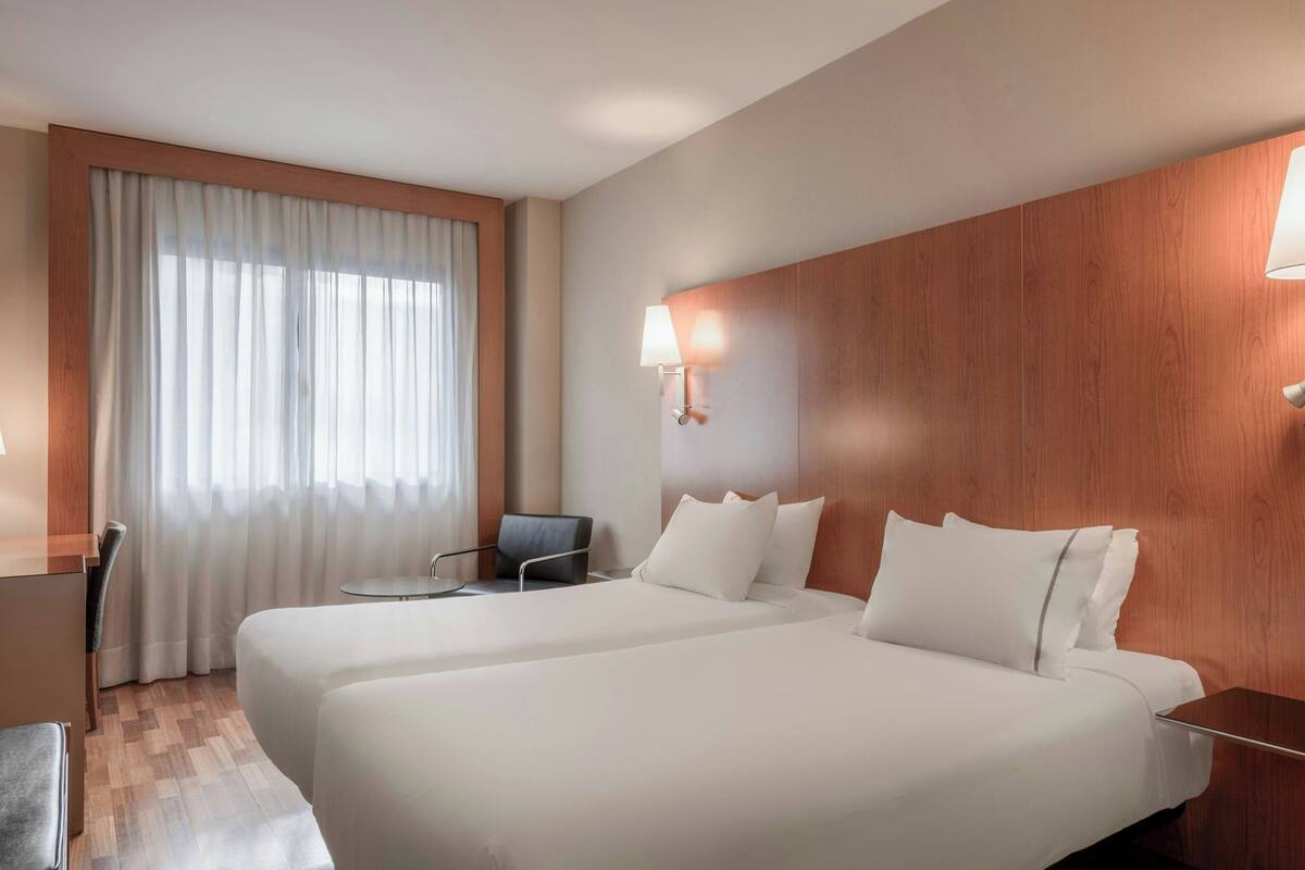 A hotel room with two beds, a desk, a chair, and a large window with curtains.