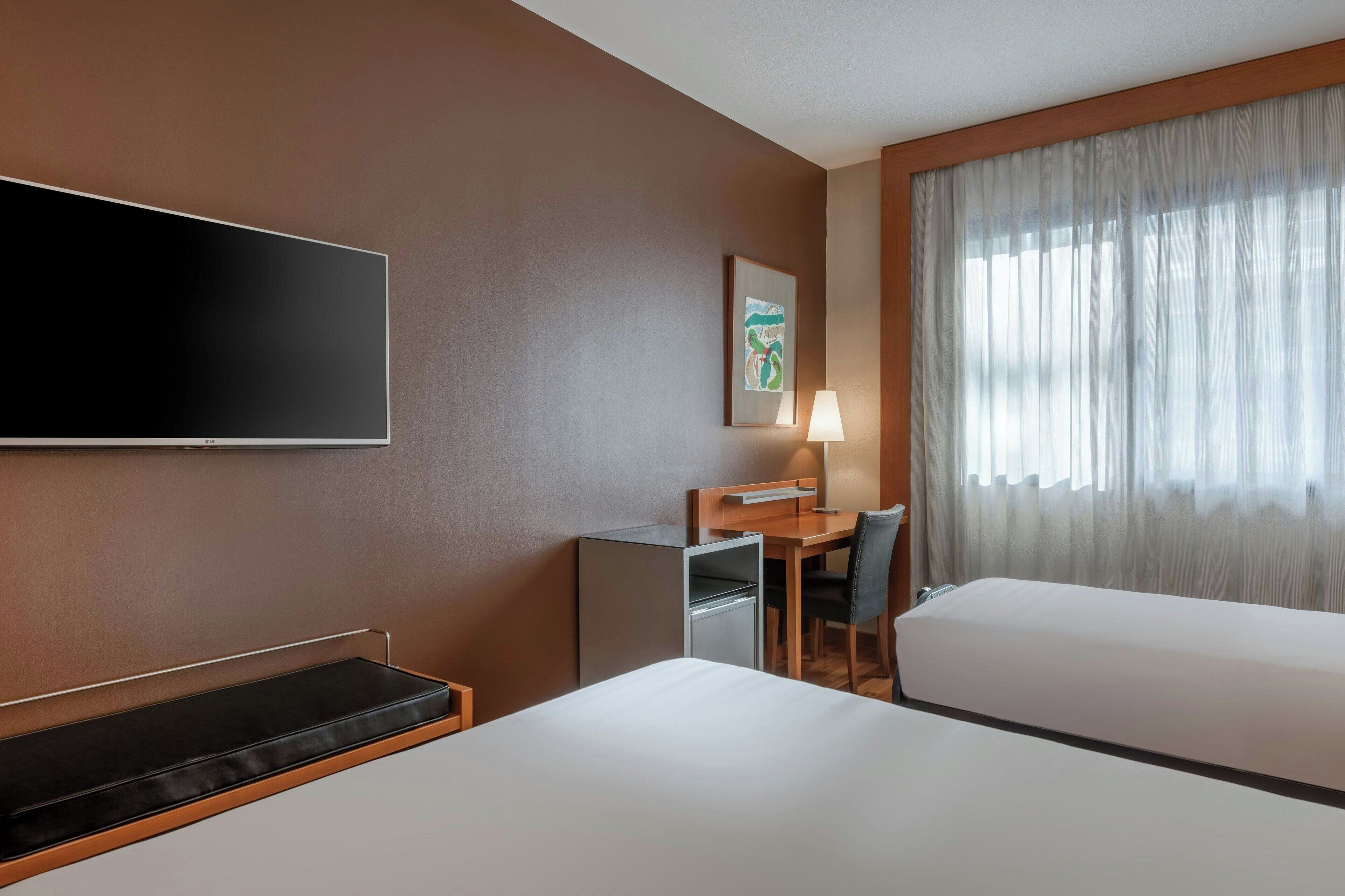 Een hotelkamer met een bed, een bureau met stoel, een televisie en een raam met gordijnen.