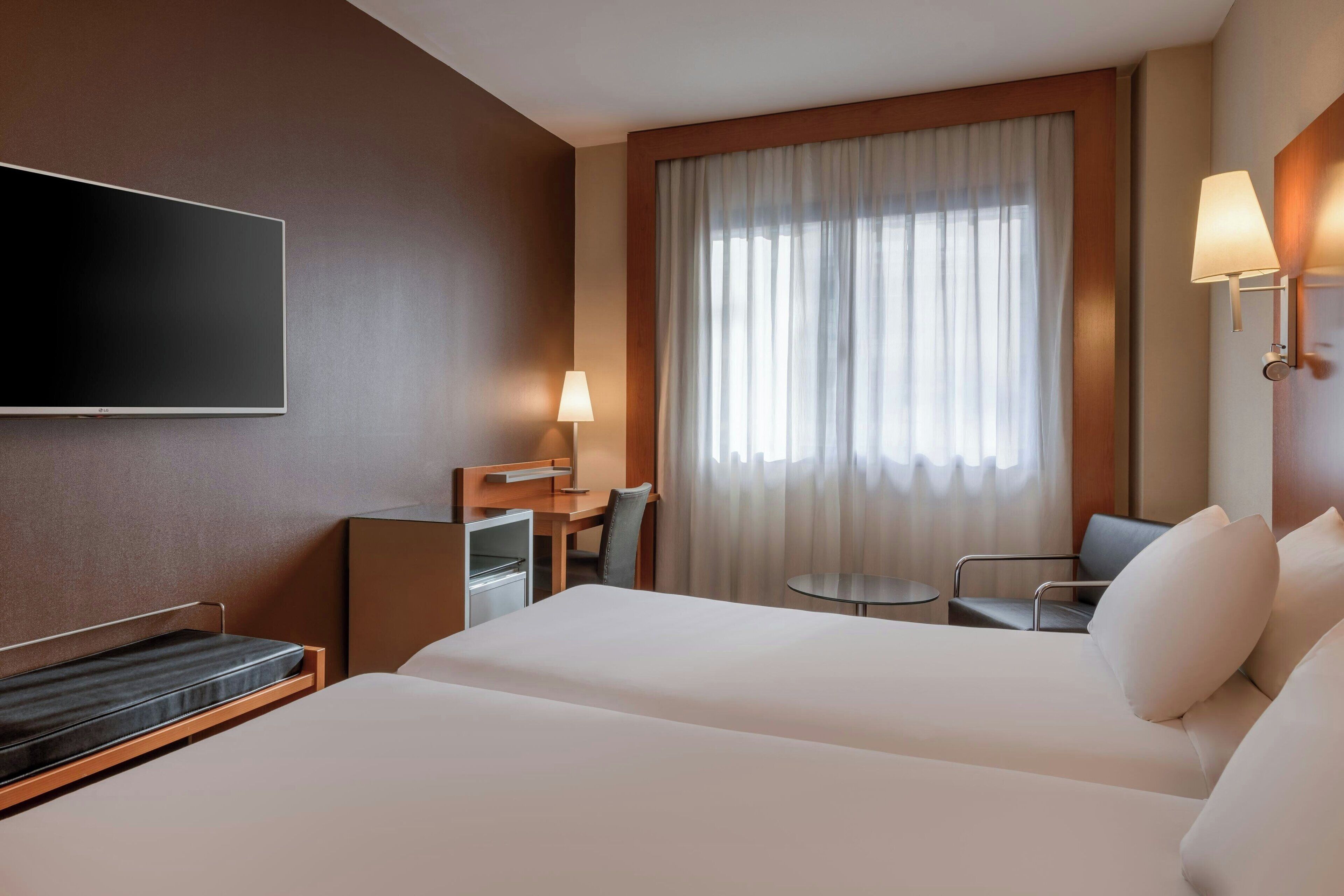 Habitación de hotel con una cama grande, un escritorio con silla, un televisor montado en la pared y una ventana con cortinas.
