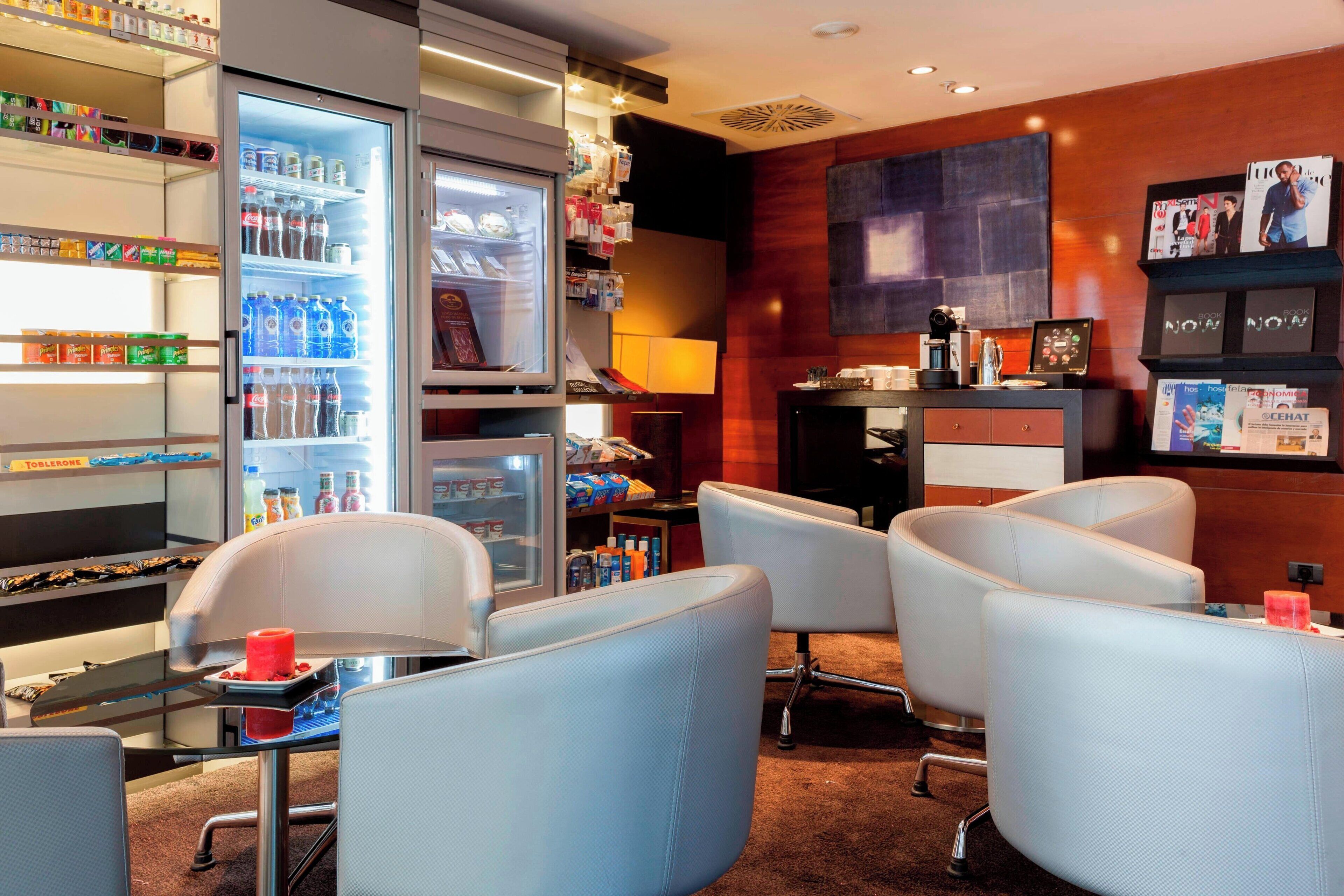 A hotel lobby with seating, a refrigerator stocked with beverages, and magazines on shelves.