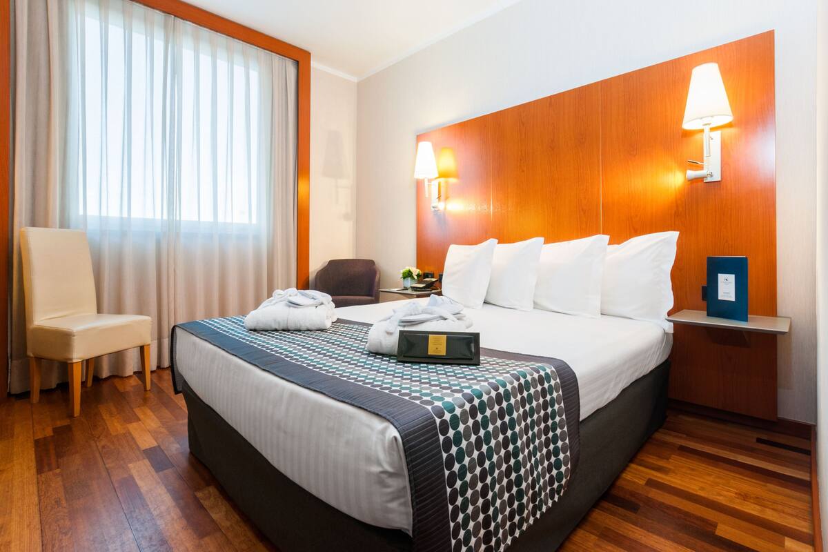 A hotel room with a large bed, a chair, a nightstand with a lamp, and a view of the outside through sheer curtains.