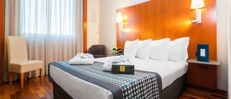 A hotel room with a large bed, a chair, a nightstand with a lamp, and a view of the outside through sheer curtains.