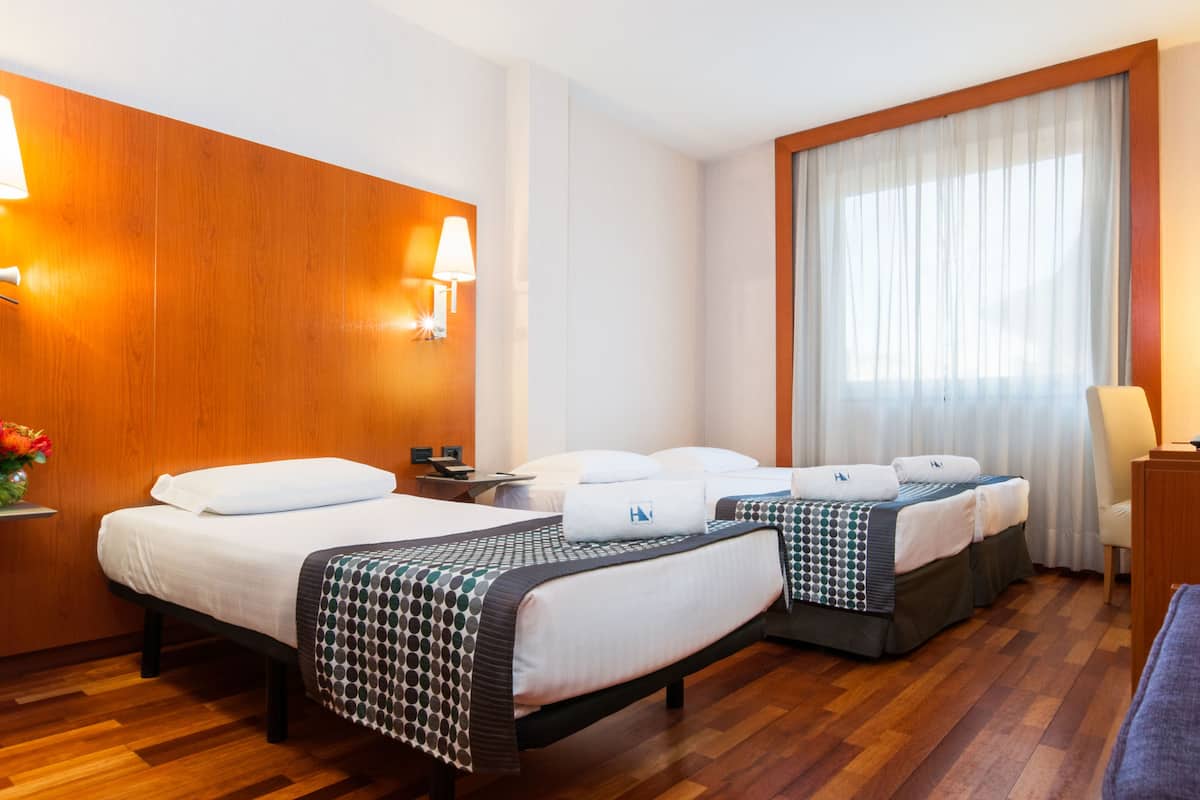 A hotel room with two beds, wooden paneling, a large window with sheer curtains, and a nightstand with a lamp.