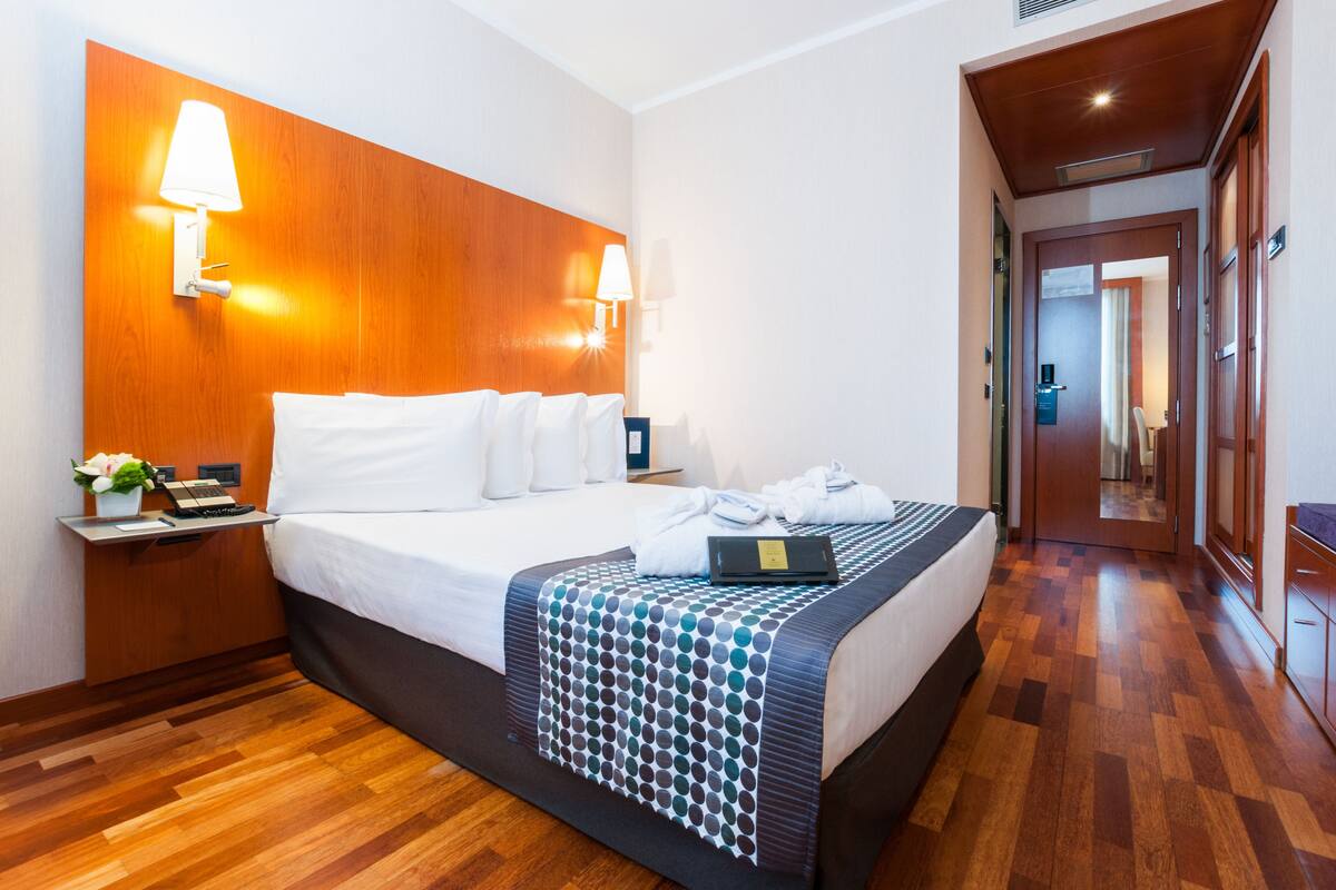 A hotel room with a wooden headboard, a bed with white linens, a bedside table with a lamp, a laptop, and a plant.