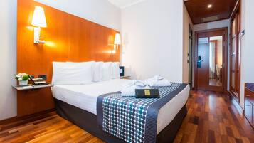 Habitación de hotel con cabecera de madera, cama con ropa blanca, mesita de noche con lámpara, una computadora portátil y una planta.